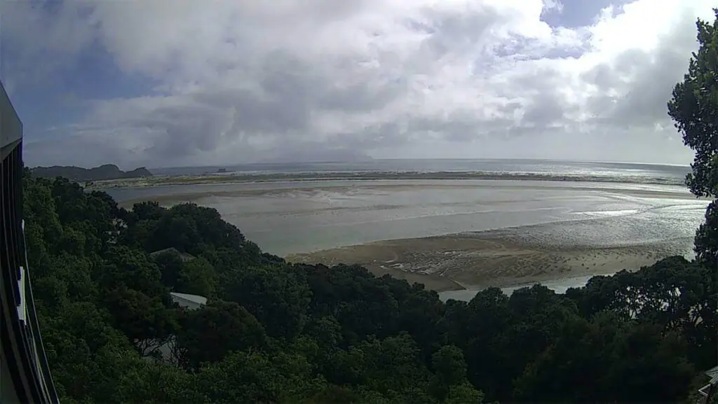 Coastal view of a tidal flat surrounded by forested hills under a partly cloudy sky.